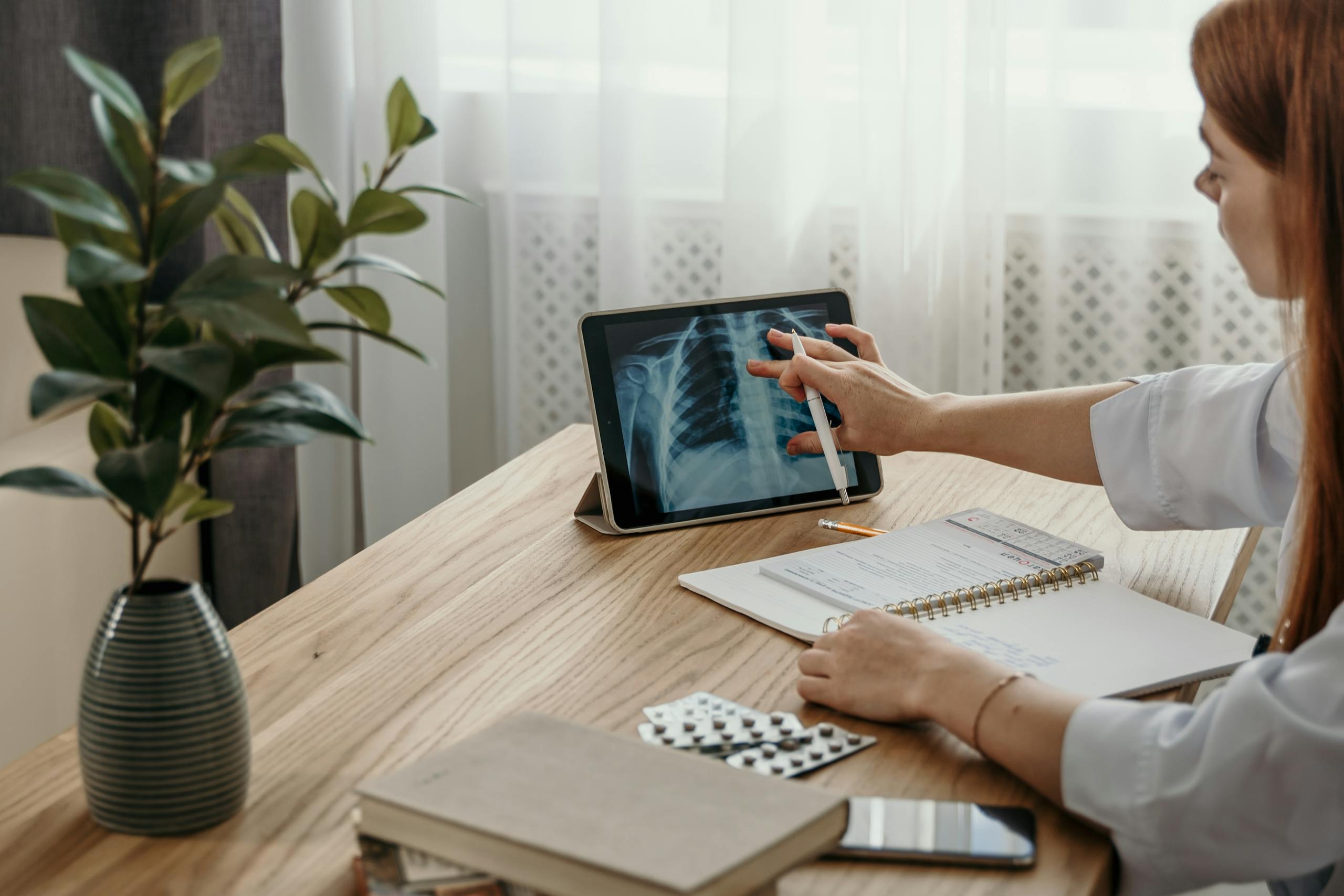 Healthcare professional examines X-ray image on tablet at desk with medical notes.